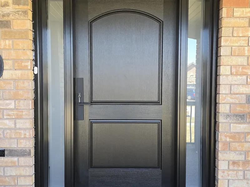 Black fiberglass entry door with an arched two-panel design and frosted glass sidelites in a tan brick wall